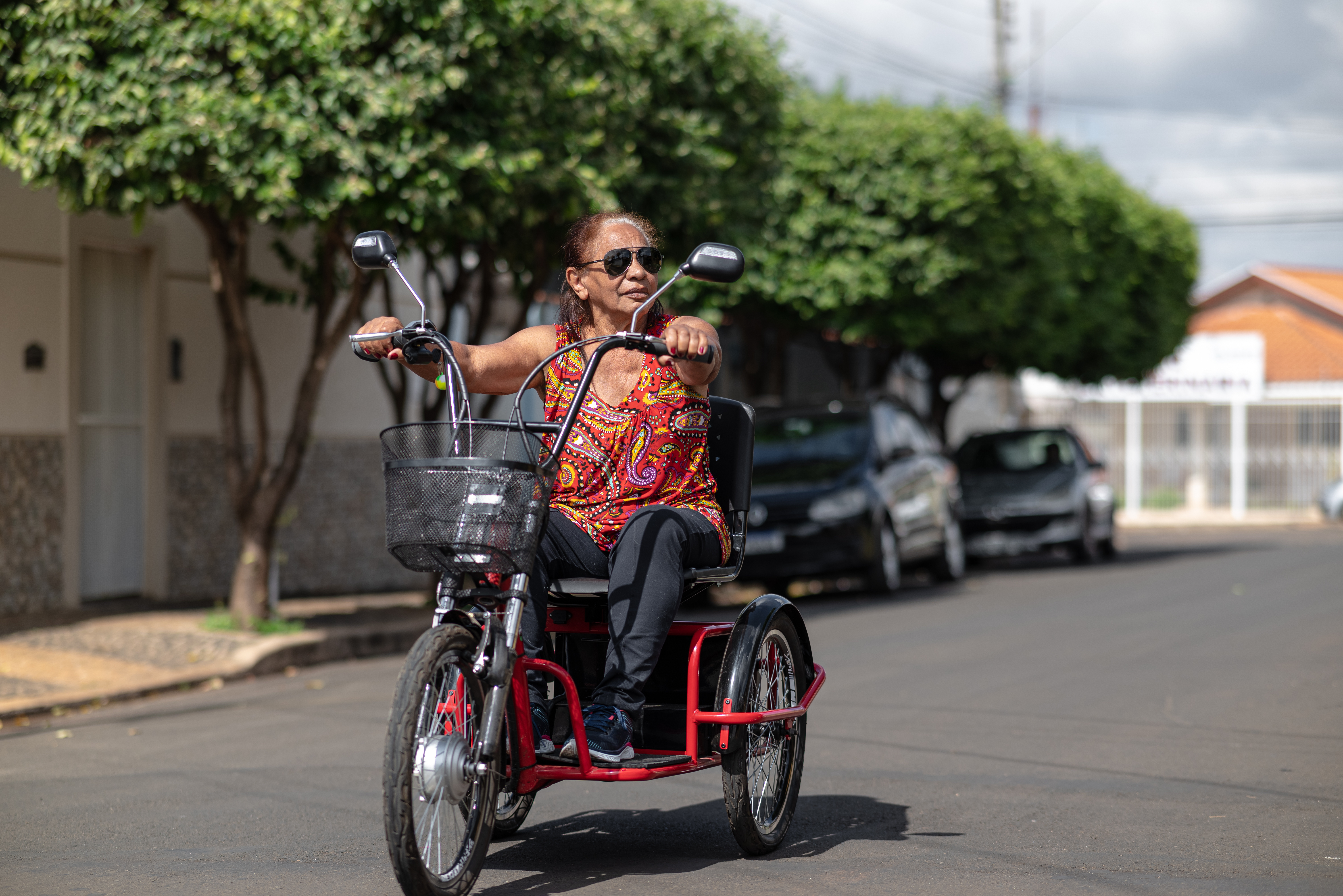 Portraits of a Black senior lady with much vitality and joy, wearing cool sunglasses and hanging out in a small city with her electric tricycle. She is riding on  calm streets with some modest and soporific houses around her, in a summer day with blue sky with clouds.